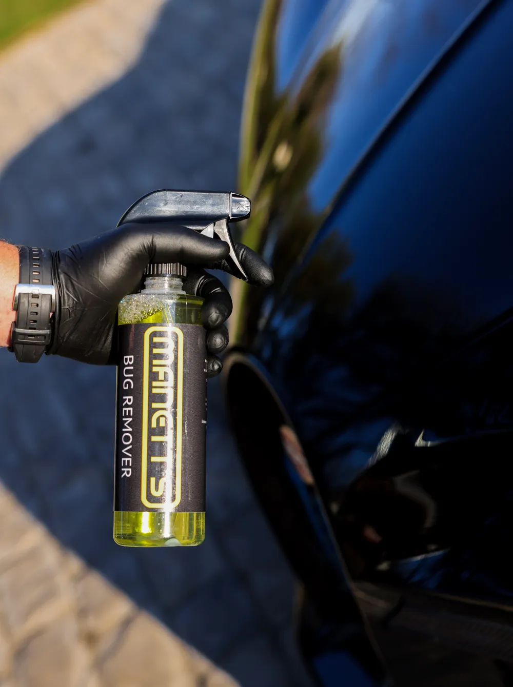Gloved detailer holding a Manetts Bug Remover bottle against the glossy black panel of a vehicle
